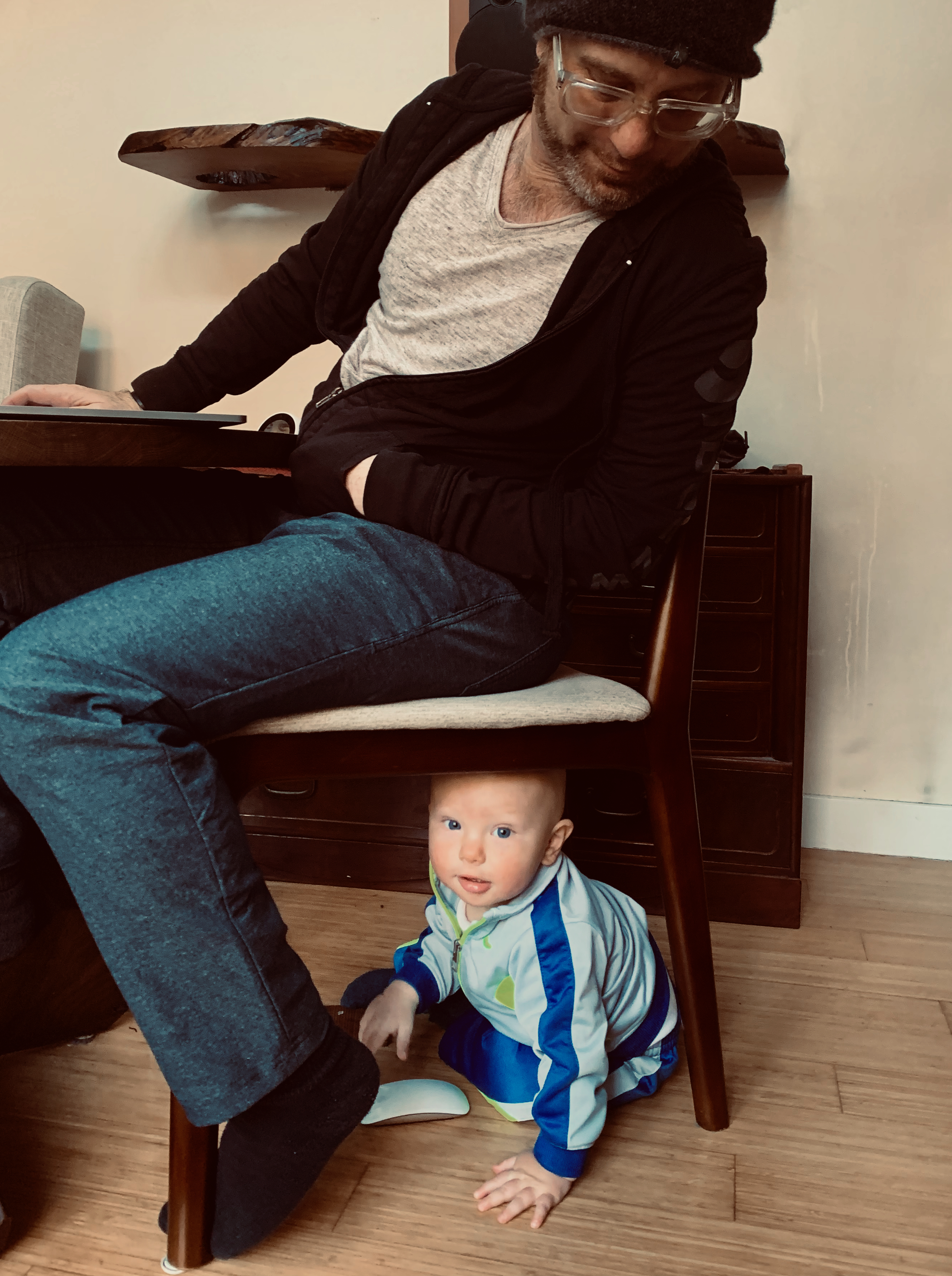 baby hiding under a chair with father sitting on top looking down
